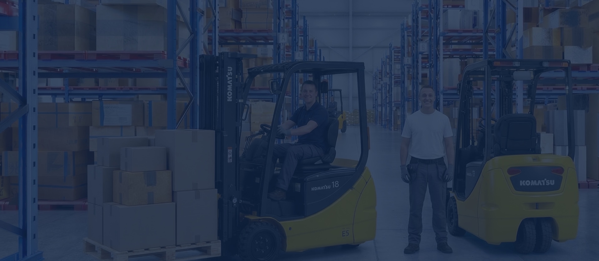 Un employé Kirmar conduit un chariot élévateur Komatsu, tandis qu’un collègue se tient entre un second chariot et les rayonnages chargés de boîtes.