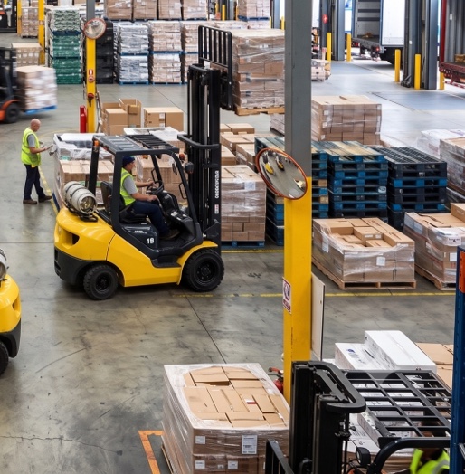 Des employés en gilets à haute visibilité conduisent des chariots élévateurs jaunes entre des palettes de marchandises dans un entrepôt.